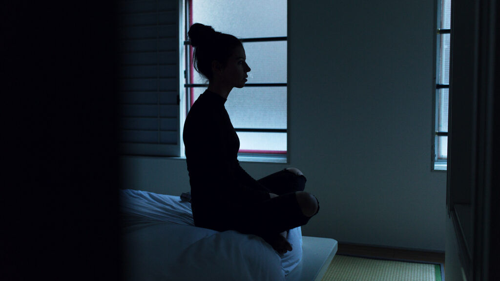 Woman sits at edge of her bed in dark room