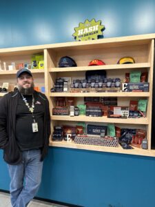 Male Budtender leaning against cannabis merchandise display case on a teal wall. 