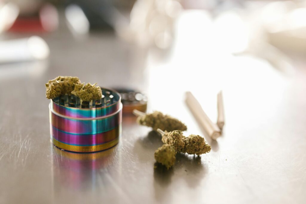 Close up of rainbow-colored weed grinder with cannabis on top of it and joints next to it on a table.