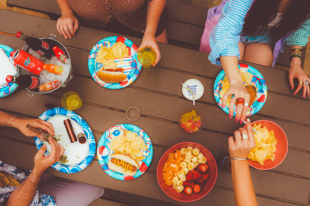 picture of three people around a picnic table from above, passing a joint and eating hot dogs