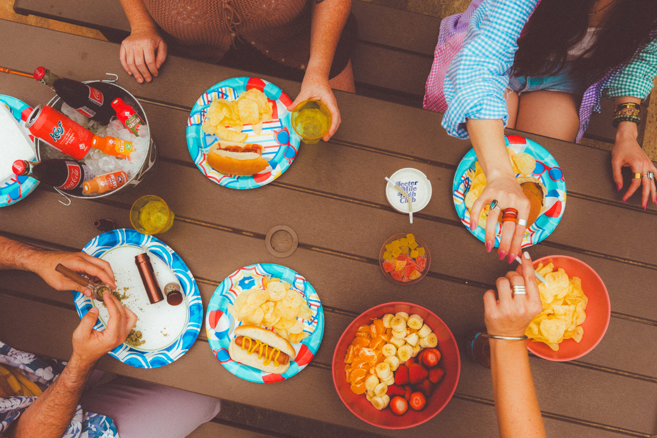 picture of three people around a picnic table from above, passing a joint and eating hot dogs