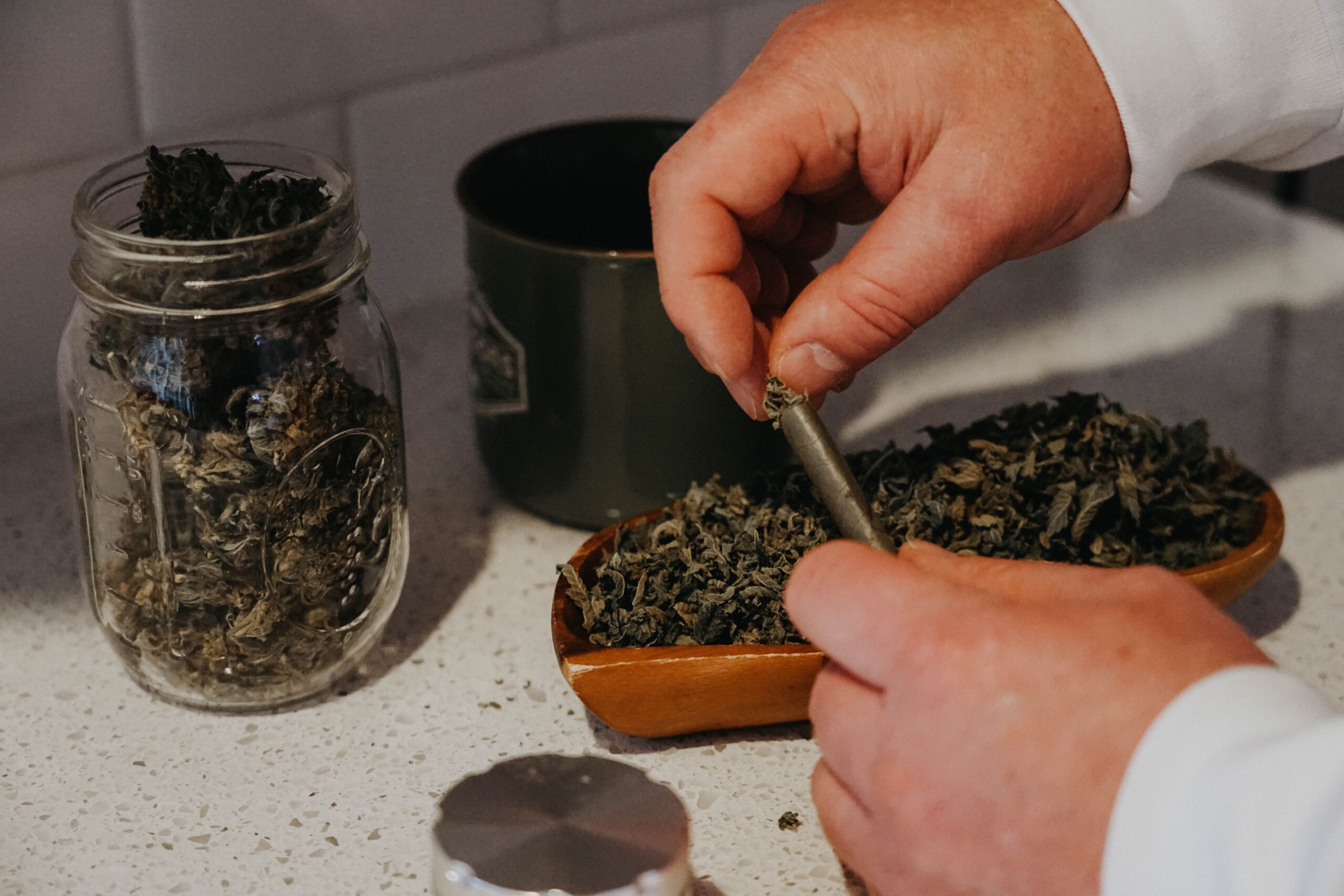cannabis flower in a jar and bowl, with a pair of hands packing a joint