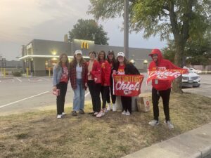 Six people wearing red and holding Chiefs flags in a McDonalds parking lot 